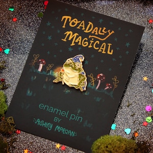 May include: A gold enamel pin featuring a green frog wearing a blue and gold wizard hat. The frog is holding a flower in its right hand. The pin is on a black card with the text "Toadally Magical" and "enamel pin by Ashley Morgan".