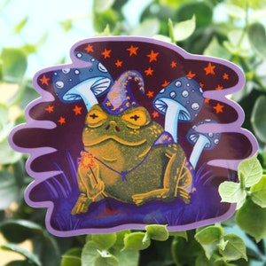 May include: A whimsical sticker featuring a green frog wearing a purple wizard hat, holding a sparkler. The frog is surrounded by three blue and white mushrooms with white dots, and a purple background with orange stars.