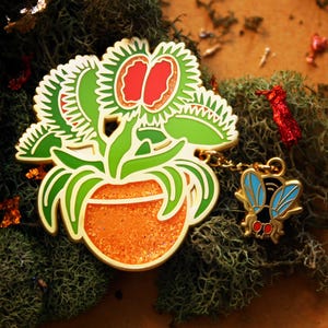 May include: A gold-toned enamel pin featuring a green Venus flytrap plant in an orange glitter pot. The plant has red interior traps with jagged edges. A small gold-toned charm of a fly with blue wings and red eyes hangs from the main pin by a chain.