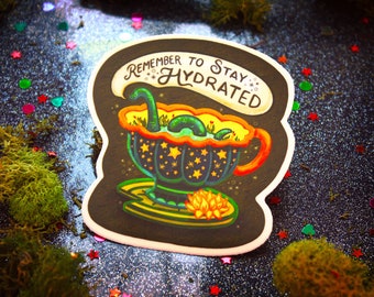 May include: A black and green sticker with a cartoon illustration of a green sea monster in a teacup. The text "Remember to Stay Hydrated" is written in a whimsical font above the monster.