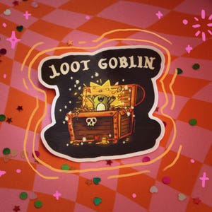 Puede incluir: Una pegatina con las palabras "LOOT GOBLIN" sobre una ilustración de un cofre del tesoro. El cofre está abierto, revelando monedas de oro, una rana y luces brillantes. La pegatina tiene un fondo negro con un borde blanco, sobre un fondo a cuadros rosa y naranja.