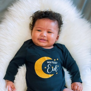 May include: A baby wearing a black long-sleeved onesie with a gold glitter crescent moon and stars. The onesie has the text "My First Eid" in white.