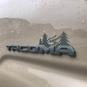 以下が含まれることがあります： ベージュのトラックのクローズアップ。黒い「TACOMA」のエンブレムと灰色の山のシーンのステッカーが貼られています。トラックは水滴で覆われています。