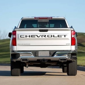 May include: A white Chevrolet Silverado pickup truck with a black tailgate and the word "CHEVROLET" in black lettering on the tailgate. The truck has a black bumper and black wheels.