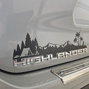 Peut inclure: Un SUV gris avec un autocollant noir représentant une chaîne de montagnes, des arbres, une tente et le mot "HIGHLANDER" à l'arrière.