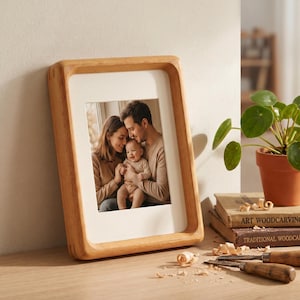 May include: A rectangular light-toned wooden picture frame with rounded inner corners containing a photograph of two adults holding a baby. The frame has a thick, smooth border and a white mat surrounding the central portrait image.