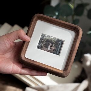 May include: A square wooden photo frame with a white mount and a small photograph. The frame is made of dark wood and has rounded corners. The photograph shows a person in a garden setting.