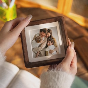 Könnte beinhalten: Ein quadratischer Bilderrahmen aus Holz mit dunkelbraunem Finish und weißem Innenrand. Der Rahmen enthält ein Foto einer Familie, bestehend aus zwei Erwachsenen und zwei Kindern, die sich umarmen. Der Rahmen wird von zwei Händen gehalten.