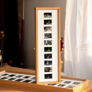 Può includere: Una cornice portafoto verticale in legno chiaro, contenente otto piccole foto rettangolari. La cornice ha un passe-partout bianco attorno a ogni foto. Le foto sembrano ritrarre persone in vari ambienti esterni. Una seconda cornice simile è visibile in primo piano.