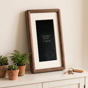 a wooden frame with a black chalkboard-style message on it, placed on a white surface next to three potted plants and a set of keys.