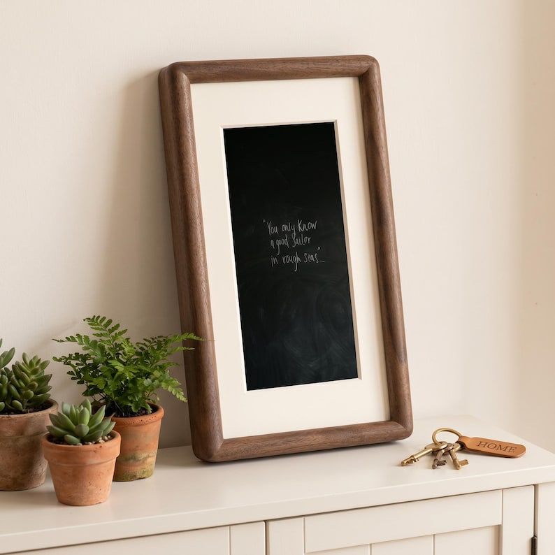 a wooden frame with a black chalkboard-style message on it, placed on a white surface next to three potted plants and a set of keys.