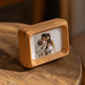 Könnte beinhalten: Ein rechteckiger Bilderrahmen aus Holz mit abgerundeten Kanten und einer natürlichen hellbraunen Maserung. Der Rahmen enthält ein Foto einer vierköpfigen Familie, bestehend aus zwei Erwachsenen und zwei Kindern, die sich eng umarmen.