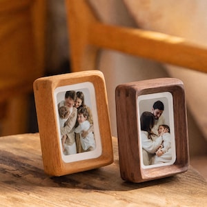 Peut inclure: Deux cadres photo rectangulaires en bois aux bords arrondis, l'un en bois clair et l'autre en bois brun foncé. Chaque cadre contient une photographie verticale d'une famille, avec une bordure blanche autour de l'image à l'intérieur du cadre.