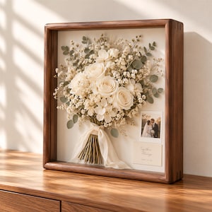 May include: A square wooden shadow box frame containing a preserved bouquet of white roses, baby's breath, and eucalyptus tied with a cream ribbon. A small photograph of a couple and a card with the text Together Always 2024.10.26 are included.