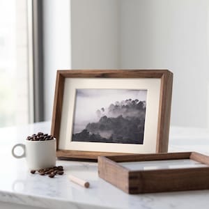 Könnte beinhalten: Ein rechteckiger Bilderrahmen aus Holz mit cremefarbenem Passepartout, der ein Schwarz-Weiß-Foto eines nebligen Waldes enthält. Daneben steht eine kleine weiße Keramiktasse mit dunklen Kaffeebohnen sowie ein zweiter, leerer Holzrahmen.