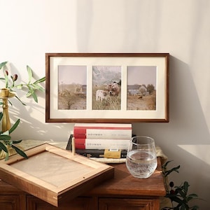 May include: A rectangular wooden picture frame with a cream-colored mat featuring three vertical openings. The frame displays a triptych of rural landscape photographs showing cows in a field with mountains in the background and a farmhouse.