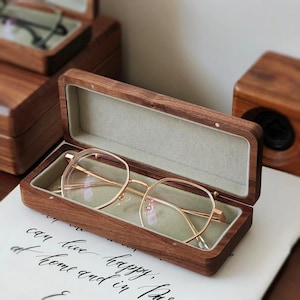 Könnte beinhalten: Eine Brille mit goldfarbenem Rahmen liegt in einem offenen Brillenetui aus Holz. Das Etui ist mit hellgrauem Stoff ausgekleidet. Die Brille hat eine moderne, geometrische Form. Das Etui ist aus dunklem Holz.