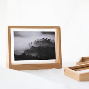 May include: A wooden picture frame with a black and white photo of a foggy forest. The frame is standing on a white surface.