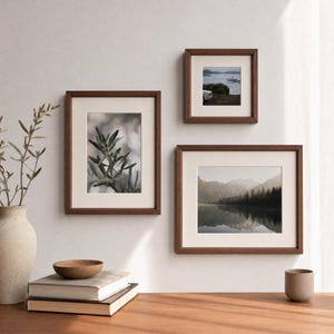 May include: Three dark wood picture frames with off-white matting hanging on a wall. The frames contain prints of an olive branch, a coastal scene, and a mountain lake. A ceramic vase, a wooden bowl, and two stacked books sit on a surface.