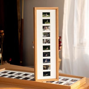 Könnte beinhalten: Ein hoher, rechteckiger Holzfotorahmen mit weißem Passepartout, der acht kleine Fotos enthält. Der Rahmen besteht aus hellfarbigem Holz. Ein zweiter, ähnlicher Rahmen liegt flach im Vordergrund. Der Rahmen ist etwa 60 cm hoch.