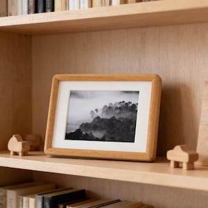 May include: A wooden picture frame with a black and white landscape photo of a forest in the fog. The frame is light brown and sits on a wooden shelf with other decorative wooden objects and books.
