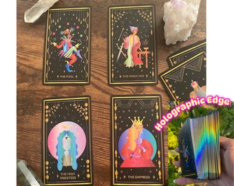 May include: Four tarot cards with gold accents and holographic edges. The cards are laid out on a wooden surface. The cards depict The Fool, The Magician, The High Priestess, and The Empress.