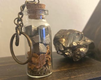 May include: A small glass bottle with a cork stopper and a metal chain attached. The bottle is filled with small silver metal pieces and brown dried herbs. The bottle is sitting on a wooden surface.
