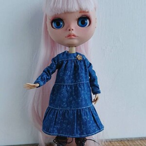 以下が含まれることがあります： 長いピンクの髪をした人形が、花柄の青いデニムのドレスを着ています。人形は茶色のブーツを履いており、木製の表面に立っています。