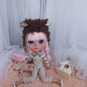 Op de afbeelding: Een op maat gemaakte Blythe-pop met roze haar en blauwe ogen, gekleed in een bruine berenmuts en een geruite overall. De pop houdt een kleine teddybeer vast en poseert met een houten vogelhuisje.
