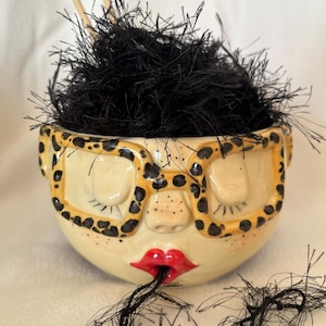 May include: A whimsical ceramic bowl with a face design, featuring closed eyes, a nose, and red lips. The bowl has a yellow and black leopard print pattern around the rim, resembling glasses. The bowl is filled with black, fluffy material.