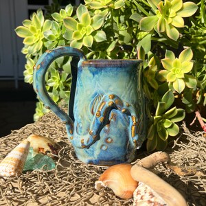 May include: A ceramic mug with a blue and gold octopus design. The mug has a textured surface and a large handle. The background includes seashells, driftwood, and green plants.