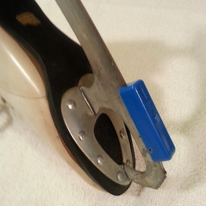 May include: Close-up of a white figure skate with a black boot and a silver blade. The blade has a blue plastic attachment on the toe.