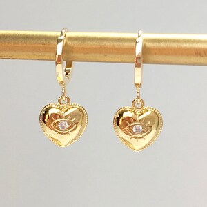 Puede incluir: Un par de pendientes de aro dorados con un dije en forma de corazón que presenta un diseño de ojo malvado con un strass transparente en el centro.