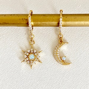 May include: A pair of gold hoop earrings with a dangling star and crescent moon charm. Each charm is adorned with sparkling clear crystals and a small opal stone in the centre.