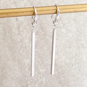 May include: A pair of silver dangle earrings with a small clear crystal at the top of each earring and a long, thin silver bar hanging below.