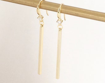 May include: A pair of gold dangle earrings with a small clear rhinestone at the top of each earring. The earrings feature a long, straight, gold bar that hangs from the rhinestone.