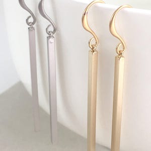 May include: Two pairs of drop earrings featuring long, slender rectangular bars suspended from curved ear wires. One pair is finished in a cool silver tone, while the other pair is finished in a warm gold tone, both with a smooth, matte surface.
