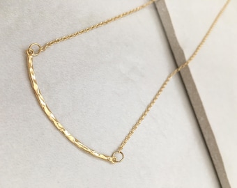May include: A delicate gold-colored necklace featuring a curved bar pendant with a hammered texture. The pendant is connected to a fine chain, creating a minimalist and elegant design. The necklace is displayed on a neutral background.