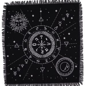 May include: Black square textile featuring a white celestial design. The design includes a smiling sun, moon with a face, zodiac symbols, stars, and geometric shapes. The edges are fringed.