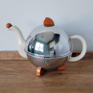 以下が含まれることがあります： 光沢のあるクロームボディ、茶色のボールチップが付いたクリーム色の注ぎ口、同色のクリーム色のハンドル、そして上部のフィン、ベースの脚、金属球の前面にある小さなラッチを含むオレンジ色のアクセントが特徴の球形のティーポット。