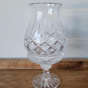 May include: A clear glass hurricane candle holder featuring a bulbous body with a diamond-cut pattern, a flared rim, and a pedestal base with a starburst design. The glass is transparent and reflects light through its intricate geometric facets.