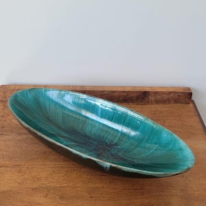 May include: An elongated oval ceramic bowl featuring a glossy glaze with a deep teal and emerald green colour gradient. The interior surface displays a radiating brushstroke pattern that transitions into a dark, near-black centre point.