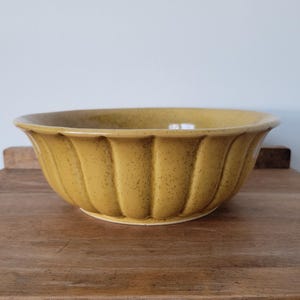 May include: A round, mustard yellow ceramic bowl with a speckled finish and a fluted, scalloped exterior design. The bowl features a wide, smooth rim and a curved body that tapers down to a circular base, showcasing a consistent glaze throughout.