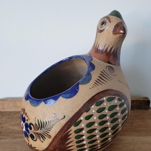 May include: A tan ceramic bird-shaped planter with a hollow opening on its back. The piece features hand-painted details in blue, green, and brown, including floral motifs on the front and a pattern of green teardrop shapes with white dots on the side.