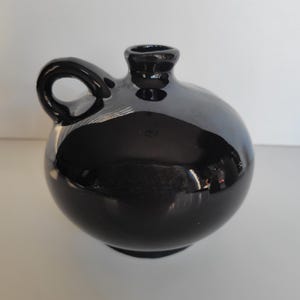 May include: A small, round, black ceramic jug with a glossy finish. The vessel features a short, narrow neck with a flared rim and a single, curved handle attached to the side. The surface is smooth and reflective, showing light highlights.