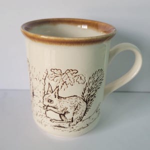 以下が含まれることがあります： 木の枝にとまったリスの茶色の線画が描かれたクリーム色の陶器製マグカップ。マグカップは滑らかな円筒形の本体に湾曲したハンドルが付いており、上部の縁には特徴的な茶色の釉薬が施されています。