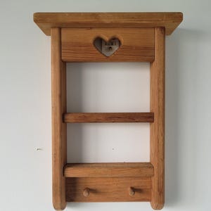 May include: A wall-mounted wooden shelf featuring a natural light brown finish, a top ledge, a middle shelf, and a bottom section with two small wooden pegs. The upper panel includes a cutout in the shape of a heart, revealing the wall behind it.