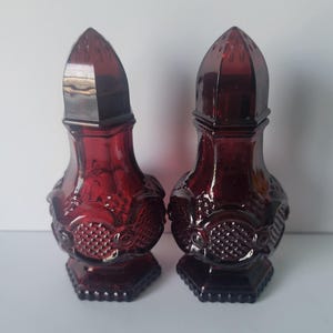 May include: A pair of deep red glass salt and pepper shakers featuring a faceted, pressed pattern with a diamond-like texture on the rounded bodies. Each shaker has a pointed, dome-shaped lid with small holes and sits on a hexagonal base.