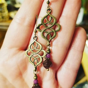 May include: A pair of dangle earrings with a gold-toned, intricate, knotted design and a dark purple gemstone at the bottom of each earring.
