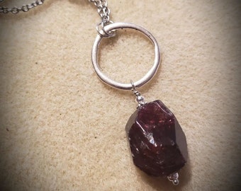 May include: A silver chain necklace with a silver ring pendant and a dark red gemstone. The gemstone is a rough, natural shape.